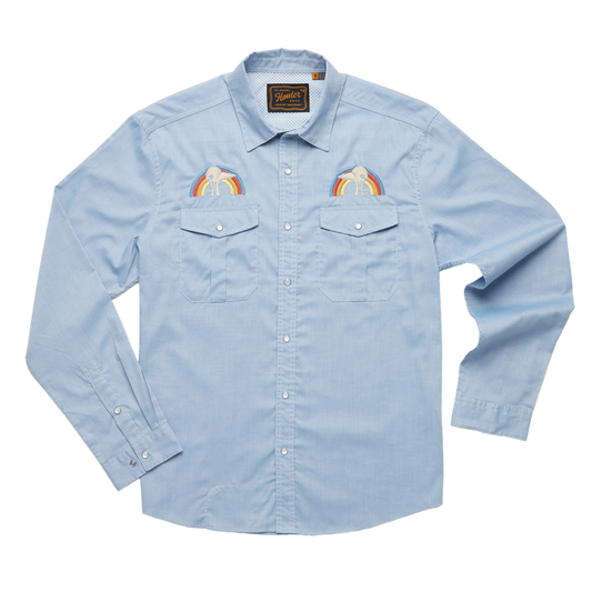 A long-sleeved blue shirt with a rainbow-colored bird embroidery on the chest and a text logo on the back shoulder. It features pearl snaps and a sunglass cleaning microfiber at the hem.